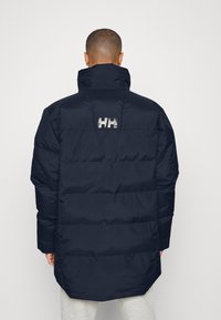 Marineblå pufferjakke med vandrette quiltede sektioner, høj krave og et reflekterende logo på ryggen. Har et langt design og en glat tekstur.