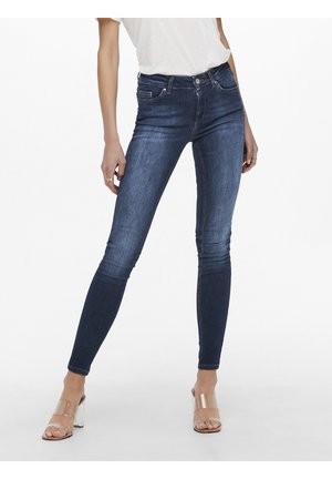 Kobieta w ciemnoniebieskich obcisłych jeansach i wysokich sandałach z przezroczystymi paskami, stojąca z lekko rozstawionymi nogami na białym tle.