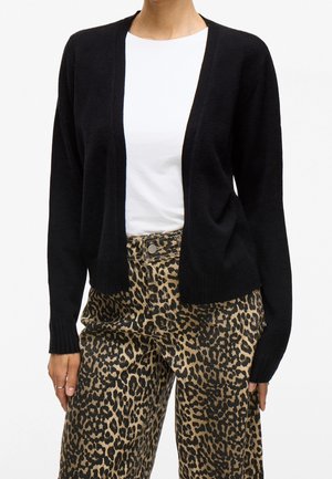 Femme portant un cardigan noir, une chemise blanche et un pantalon à imprimé léopard, debout avec les mains détendues le long du corps sur fond blanc.