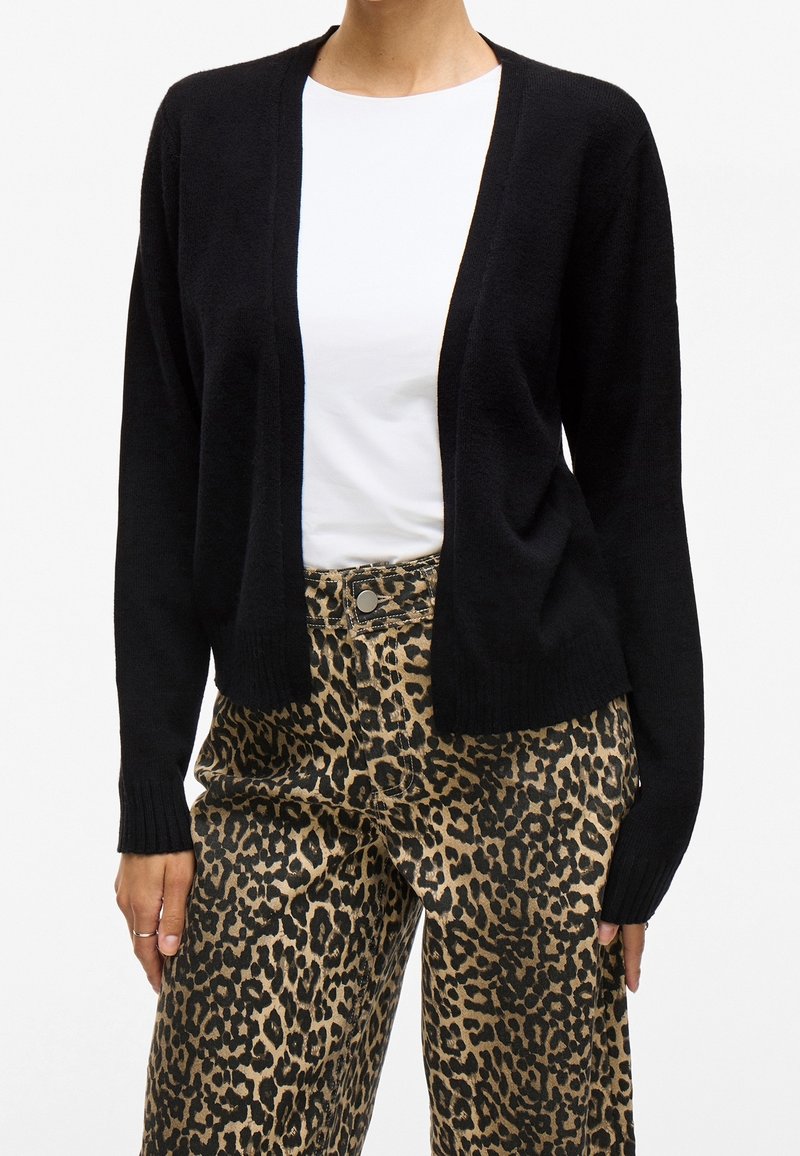 Femme portant un cardigan noir, une chemise blanche et un pantalon à imprimé léopard, debout avec les mains détendues le long du corps sur fond blanc.