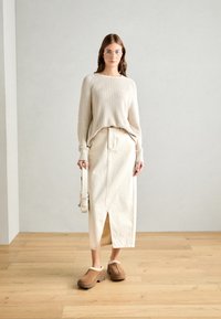 Jersey beige, falda de rayas crema, zuecos marrones sin tacón con forro de piel, y un pequeño bolso cruzado, todo dispuesto sobre un fondo neutro.