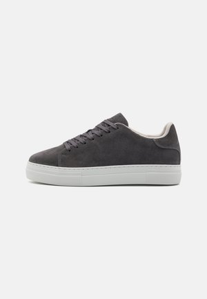 Sneaker en suède gris foncé avec un bout rond, des lacets plats et une semelle en caoutchouc blanche. Texture lisse avec des détails de couture minimaux et un rembourrage au niveau de la cheville.