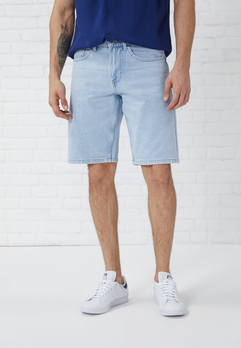 Light blue denim shorts with a regular fit, straight leg, and top-stitched hem. Paired with white sneakers and a navy button-up shirt.
