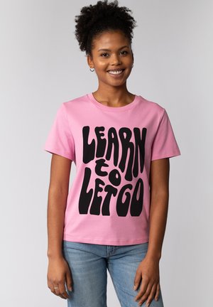 Femme souriante aux cheveux bouclés portant un T-shirt rose avec l'inscription en lettres noires épaisses "APPRENDRE À LÂCHER PRISE", associée à un jean bleu, debout devant un fond gris.