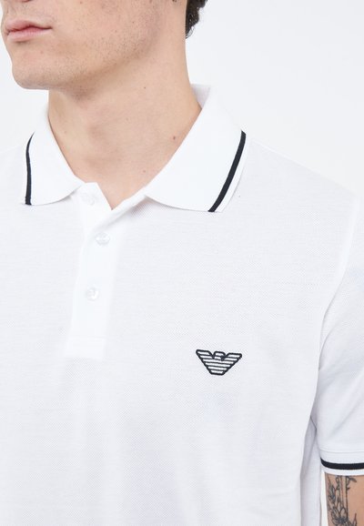 Mężczyzna ubrany w biały polo z czarnym wykończeniem przy kołnierzu i rękawach, z czarnym paskowanym logo orła na piersi.
