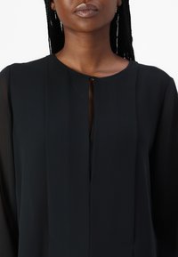 Tunic nera a maniche lunghe, con scollatura rotonda e un'apertura a forma di chiave sul petto. Tessuto leggero con una texture liscia.