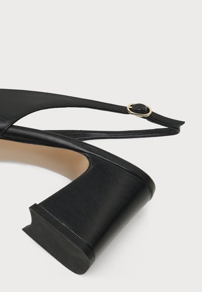 Μαύρο δερμάτινο παπούτσι slingback με χαμηλό χονδρό τακούνι και μικρή χρυσή αγκράφα στο λουράκι πάνω σε απλό ανοιχτό φόντο.