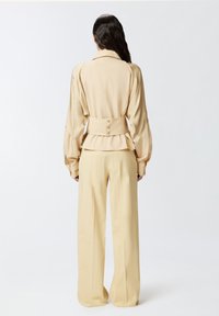 Bež košulja po mjeri s voluminoznim rukavima i uskim strukom, uparen s širokim bež hlačama, koje pokazuju glatku teksturu tkanine.