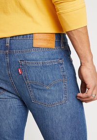 Blå denimjeans med en texturerad yta, med en läderlapp vid bakmidjan och en röd etikett på bakfickan.