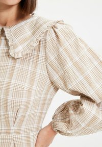 Robe à carreaux beige et blanc avec un grand col à volants et des manches longues bouffantes, portée par une personne aux cheveux bruns courts.