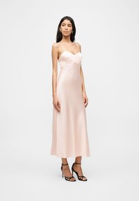 Naine kannab kahvaturoosat satiinist midi-kleiti, millel on õhukesed rihmad, ja musta kontsaga sandaale, seistes lihtsa valge tausta ees.