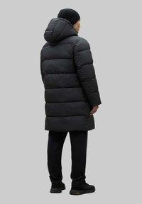 Abrigo acolchado negro con capucha, con un diseño acolchado, silueta alargada y tejido texturizado. Combinado con pantalones y calzado negros.