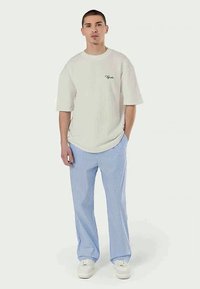 Lys grå stripete kortermet skjorte med brodert logo, sammen med løst sittende bukser i blå og hvite striper og hvite sneakers.