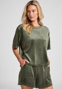 Donna dai capelli biondi che indossa un top corto in velluto verde oliva e pantaloni corti abbinati, con le mani in tasca, in piedi di fronte a uno sfondo neutro.