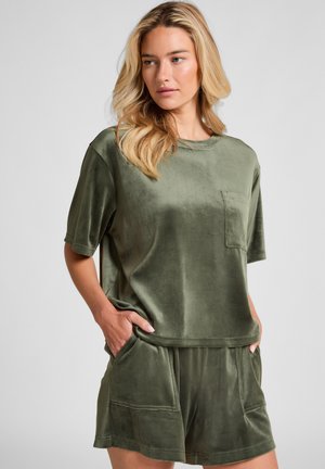 Woman with blonde hair wearing loose olive green velour short-sleeve top and matching shorts with hands in pockets, standing against plain background.