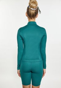 Maglietta sportiva verde a maniche lunghe con un design aderente e colletto alto, abbinata a pantaloni corti coordinati. Texture liscia con cuciture piatte.