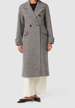 Femme portant un long manteau gris à chevrons à double boutonnage, un pantalon crème à jambes larges et des chaussures plates noires à bout pointu.