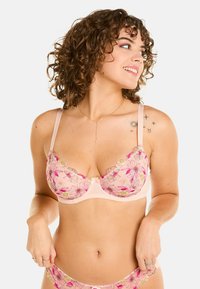Soutien-gorge en dentelle florale rose avec des motifs floraux brodés, des fines bretelles et un détail de nœud au centre. Tissu doux avec un aspect transparent.