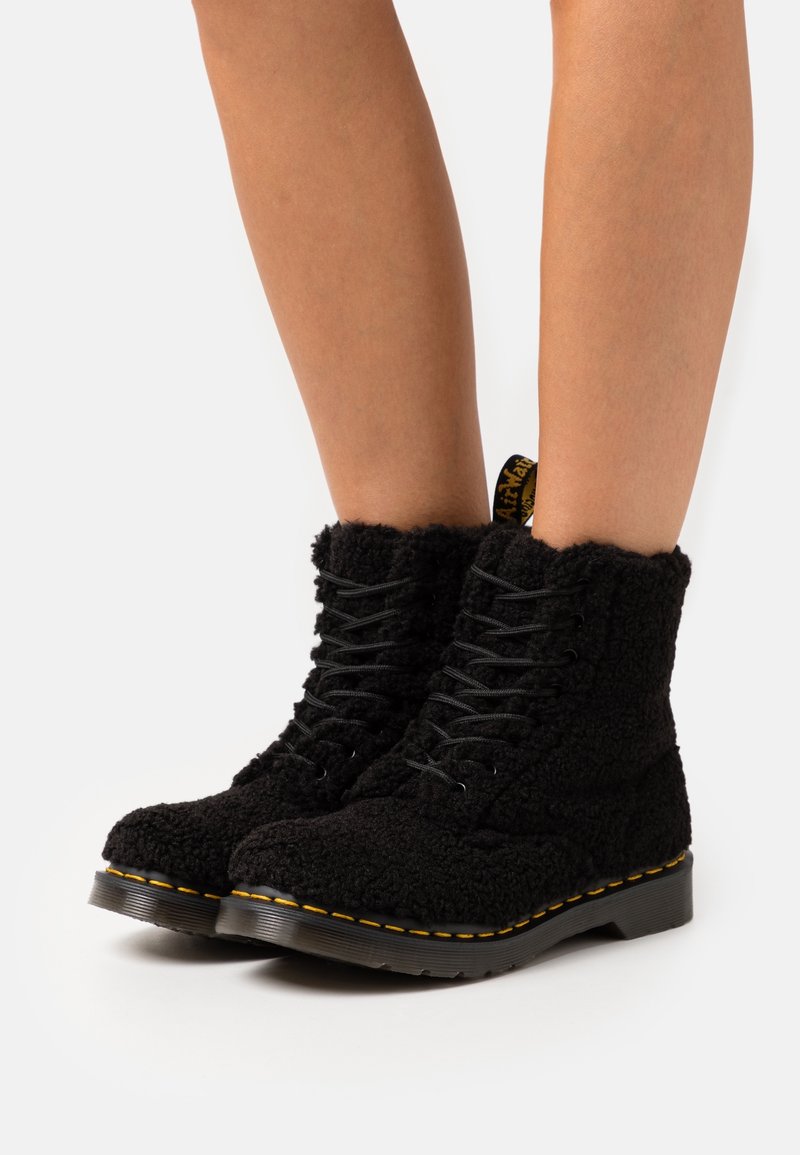 Bottines noires à lacets en tissu duveteux texturé, coutures jaunes sur la semelle, portées sur des jambes nues sur fond blanc.