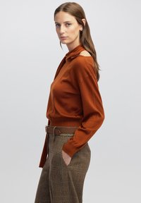Maglione lavorato a maglia in arancio ruggine con collo alto e design a spalla scoperta, abbinato a pantaloni a quadri marroni e una cintura marrone.