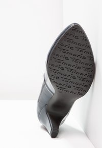 Bottine à talon haut noire à bout pointu, montrant la semelle embossée du nom de la marque "Tamaris" en répétition et la taille 37.