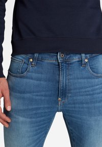 Homme portant un jean en denim bleu et un sweat-shirt bleu marine, debout avec la main gauche détendue le long du corps.