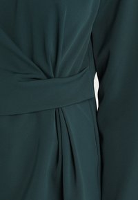 Tmavě zelené šaty z měkké látky, s zavinovacím designem, zdobené volánem a páskem v pase. Hladká textura, elegantní silueta.