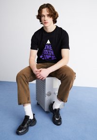 T-shirt nera con logo della montagna viola, pantaloni in corduroy beige e sneakers nere. Seduto su una scatola metallica con una superficie texturizzata.