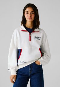 Biely fleecový pulóver s polovičným zipsom, červenými akcentmi a nápisom "Red Bull Racing". Obsahuje námornícke spodné panely a dve bočné vrecká.