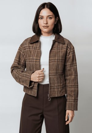 Chaqueta de cuadros marrón con cierre de cremallera y cuello ancho, con tejido texturizado. Llevada sobre una camiseta blanca y combinada con pantalones oscuros.