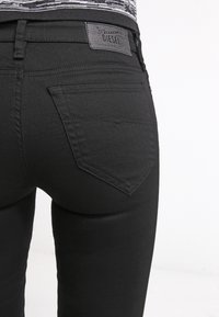 Vaqueros de mezclilla negros con un diseño ajustado, que muestran un bolsillo trasero visible y una etiqueta de cuero con la marca en la cinturilla. Textura suave.