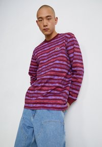 Long-sleeve top in burgundy with blue and purple horizontal stripes; features a round neckline and small embroidered logo on the chest.