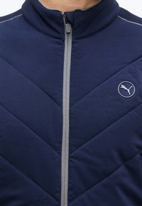 Veste matelassée bleu marine avec fermeture éclair grise et logo Puma sur la poitrine, portée par une personne dont le cou est visible.
