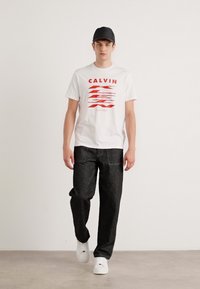 Biely bavlnený tričko s červenou abstraktnou grafikou a textom "CALVIN KLEIN", skombinované s tmavými džínsami a bielymi teniskami.