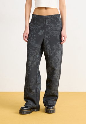 Personne portant un pantalon noir ample avec des motifs floraux subtils et des bottes noires épaisses, debout sur un sol jaune moutarde devant un mur uni.