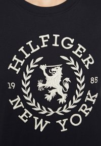 Černé bavlněné tričko s bílým vyšitým textem "HILFIGER" a "NEW YORK", s erbem lva a vavřínovými listy. Rok '1985' je zahrnut.
