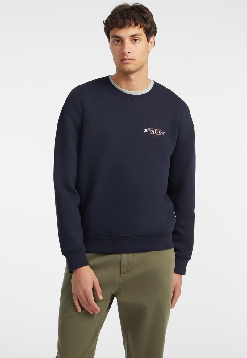 Sweatshirt em fleece azul-marinho com decote redondo ribado e punhos. Apresenta um logótipo bordado em contraste no peito. Combinado com calças verde-oliva.