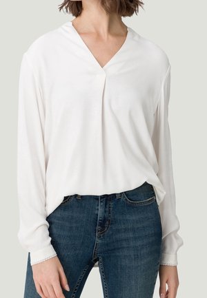 Femme portant un chemisier blanc à manches longues et col en V, rentré dans un jean en denim bleu, debout devant un fond uni.