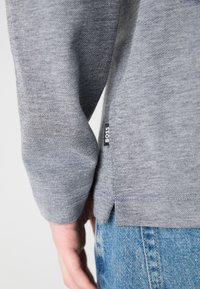 T-shirt cinza de manga longa com um tecido texturizado, apresentando uma bainha com fenda lateral e um rótulo preto "BOSS" visível perto da costura.