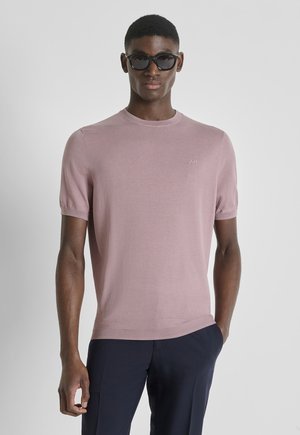 Uomo che indossa occhiali da sole neri e un maglione rosa chiaro a maniche corte con pantaloni blu scuro, in piedi con una mano in tasca.