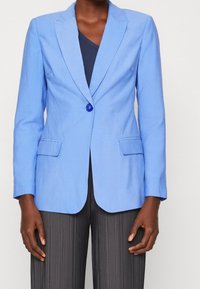 Hellblau maßgeschneiderter Blazer mit einem einzelnen Knopf, Reverskragen und zwei Fronttaschen. Über einem dunklen Oberteil getragen, kombiniert mit strukturierten dunklen Hosen.