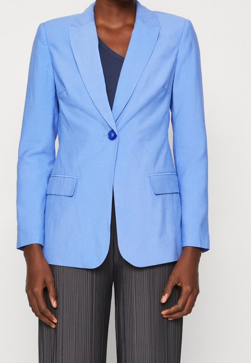 Hellblau maßgeschneiderter Blazer mit einem einzelnen Knopf, Reverskragen und zwei Fronttaschen. Über einem dunklen Oberteil getragen, kombiniert mit strukturierten dunklen Hosen.