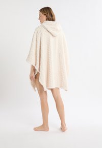 Poncho con cappuccio beige realizzato in tessuto strutturato con motivo a maglia, caratterizzato da una vestibilità ampia e un orlo che arriva poco sopra le ginocchia.