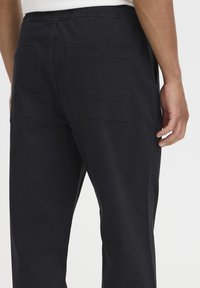 Sorte bukser med elastisk midjebånd, med to store påsydde lommer bak. Stoffet ser glatt ut med en matt finish.