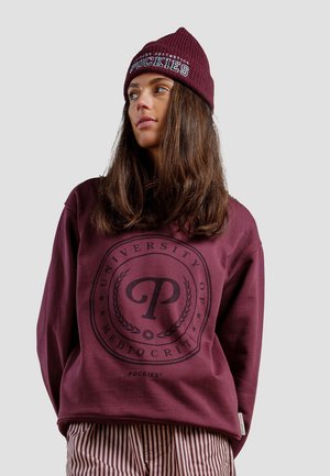 Sudadera granate con un logo circular en la parte frontal, combinada con un gorro de canalé a juego en el mismo color. Pantalones a rayas por debajo.
