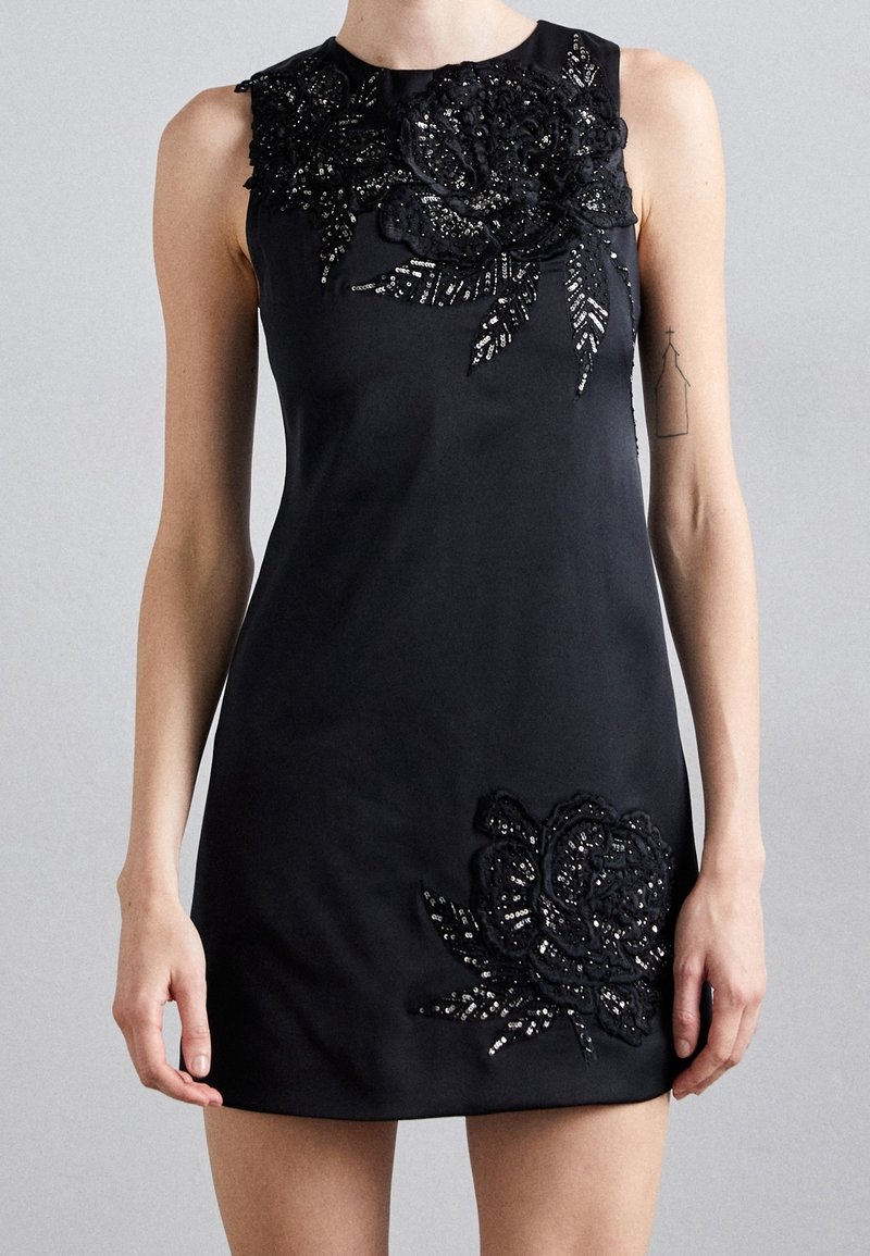 Mini-robe noire sans manches avec grandes applications florales à sequins sur la poitrine et près de l'ourlet portée par une personne debout avec un tatouage minimaliste au bras.