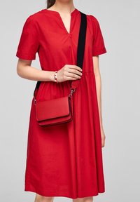 Rød bomulds kjole med V-hals og korte ærmer. En rød håndtaske med skulderrem hænger fra håndleddet. Enkel og ren design.