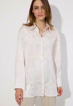 Woman wearing a long-sleeve white button-up shirt with a subtle floral pattern and beige pants, standing against a plain background.