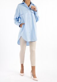 Camicia blu chiaro con bottoni e orlo arrotondato, dotata di due tasche sul petto, indossata con pantaloni beige slim-fit e décolleté color nude.