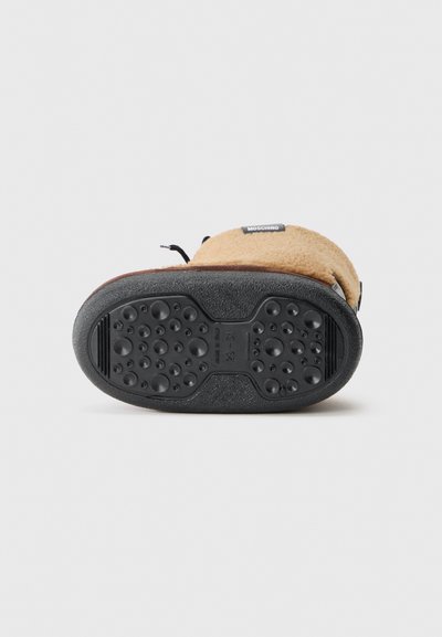 Semelle en caoutchouc noir avec motifs de grip circulaires. Dessus en polaire beige avec un accent noir, comportant une étiquette sur le côté.